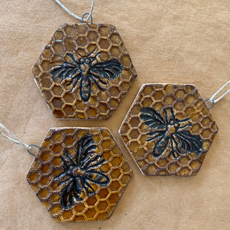 May include: Three hexagonal ceramic ornaments with a honeycomb pattern and a black bee silhouette on each. The ornaments are strung with twine.