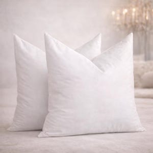 May include: Two fluffy, white pillows with pointed tops are arranged side-by-side. The pillows are made of a soft, textured fabric and appear to be filled with a down or feather material. The background is a soft, neutral color.