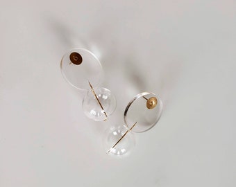 以下が含まれることがあります： 金色のアクセントが付いた透明なアクリル製のイヤリングのペア。各イヤリングは、中央に金色の円盤が付いた小さな円が、金色の棒が通っている大きな円に繋がっています。