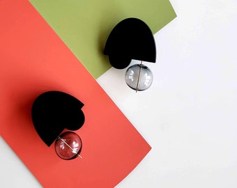 May include: Two black heart-shaped earrings with a clear glass orb hanging from each. The orb on the left earring is pink, and the orb on the right earring is gray.