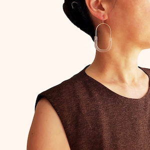 May include: A pair of gold hoop earrings with a clear acrylic oval insert. The earrings are worn by a person with brown hair and a brown sleeveless top.