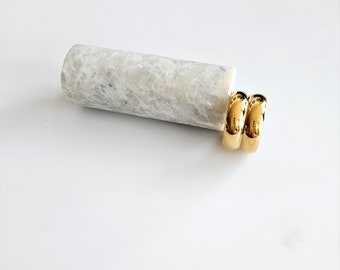 May include: Two gold-toned hoop earrings, stacked on top of each other, on a white marble surface.