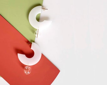 May include: A pair of white, semi-circular earrings with a small, clear glass bead hanging from each. The earrings are displayed on a green and red background.