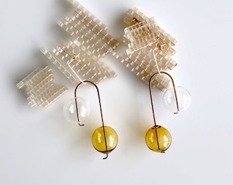 May include: A pair of modern dangle earrings with a clear glass orb and a golden-yellow glass orb suspended from a gold-colored metal hook. The earrings are paired with clear, rectangular, geometric shapes.