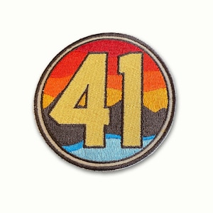 May include: A round, embroidered patch with a sunset scene and the number "41" in the center. The patch is brown with a beige border and features a blue, orange, and red sunset.