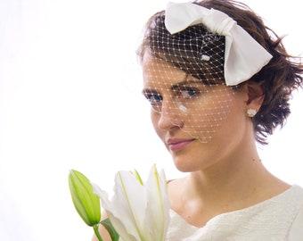 May include: A woman wearing a white bow and birdcage veil. She is holding a white lily with green leaves. The woman is wearing a white dress with a lace design. The background is white.
