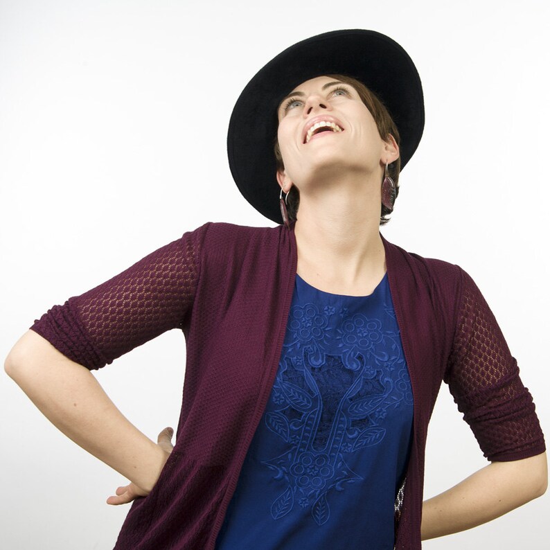 May include: A woman wearing a burgundy cardigan over a blue top with an embroidered floral design. She is wearing a black hat and has her head tilted back, looking up.