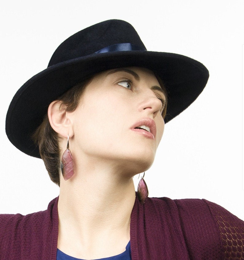 May include: A woman wearing a black fedora with a navy blue ribbon and a pair of pink and silver dangle earrings. She is wearing a burgundy sweater.