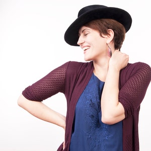 May include: A woman wearing a burgundy cardigan with a blue floral top and a black hat. She is smiling and looking to the side.