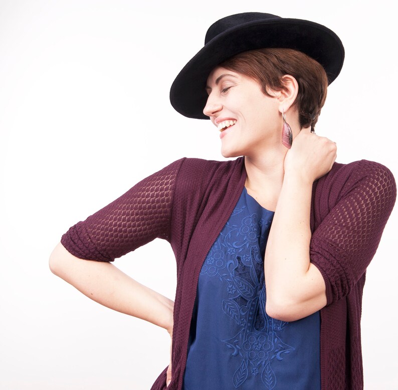 May include: A woman wearing a burgundy cardigan with a blue floral top and a black hat. She is smiling and looking to the side.