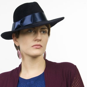 May include: A woman wearing a dark blue fedora with a navy blue ribbon and a burgundy cardigan. She is wearing pink dangle earrings.