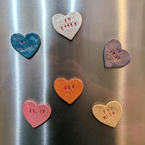 May include: A collection of heart-shaped magnets in various colors, including blue, pink, orange, and yellow. Each magnet has a different phrase in red lettering, such as "NOT YOURS" and "EW GROSS". The magnets are attached to a metallic surface.