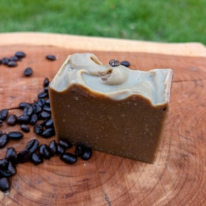 May include: A brown coffee soap bar with a swirl design and a coffee bean embedded in the top. The soap bar is sitting on a wooden surface with coffee beans scattered around it.