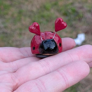 May include: A handcrafted ladybug figurine with a glossy red shell and black head. Two red heart-shaped antennae are attached with silver wire. The ladybug has small black spots and sits on a dark base. The figurine is held in a hand.