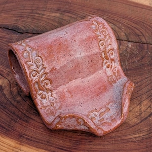 May include: A handmade, ceramic soap dish in a reddish-brown hue. The dish features an embossed floral design along the edges and a curved shape with a raised back. The surface has a speckled texture.