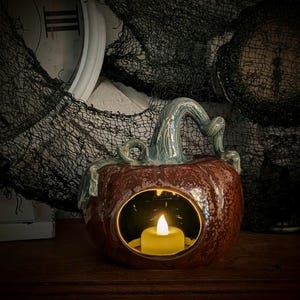 May include: A ceramic pumpkin-shaped candle holder with a teal stem and a round opening. The pumpkin is a mottled reddish-brown color and contains a lit, yellow LED candle. The background includes black fabric and a white clock.