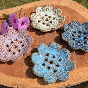 May include: Four handmade ceramic soap dishes in a flower shape, each with a unique glaze. The colors include light blue, blue, off-white, and lavender, all with a brown floral pattern. The dishes are arranged on a wooden surface.