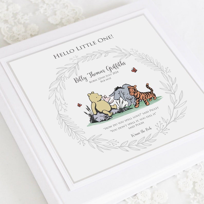 K&ouml;nnte beinhalten: Eine quadratische, wei&szlig;e Erinnerungsbox mit einem aufgedruckten grauen Blattkranz. In der Mitte sind Winnie Puuh, Ferkel, I-Aah und Tigger abgebildet. Der Text lautet: HELLO LITTLE ONE! Billy Thomas Griffiths, BORN 22ND JULY 2024, 8LB 6OZ, sowie ein Zitat.