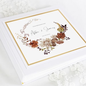 May include: White wedding album with a gold border and a watercolor illustration of fall foliage, gourds, and flowers. The text on the cover reads "Our Wedding Day Abbie & James 24.09.2022".