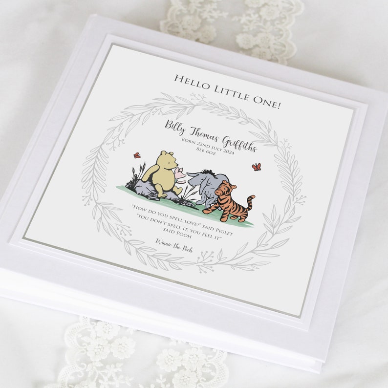 K&ouml;nnte beinhalten: Eine quadratische wei&szlig;e Erinnerungsbox mit einem Aufdruck, der den Text HELLO LITTLE ONE!, Billy Thomas Griffiths, BORN 22ND JULY 2024, 8LB 6OZ sowie eine Winnie Puuh-Illustration mit Ferkel, I-Aah und Tigger in einem grauen Blattkranz zeigt.