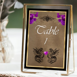Könnte beinhalten: Eine goldgerahmte Tischnummernkarte mit braunem Hintergrund. Die Karte zeigt eine Schwarzweißillustration von zwei Drachen mit einem violetten Edelstein in der Mitte. Der Text "Tisch 1" ist in weißer Kursivschrift geschrieben.