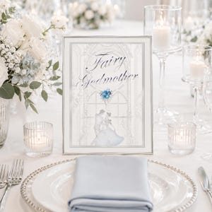 Pode incluir: Um sinal de mesa retangular com moldura prateada apresentando o texto Fairy Godmother em escrita elegante, uma ilustração de uma pedra preciosa azul e a silhueta de um casal dançando em frente a uma janela grande, em orientação vertical.
