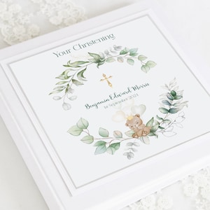 May include: A white photo album with a green and white floral design and a gold cross. The text "Your Christening" is at the top of the album. The text "Benjamin Edward Morris 1st September 2024" is below the floral design. A cartoon bear with a crown and balloons is in the center of the design.