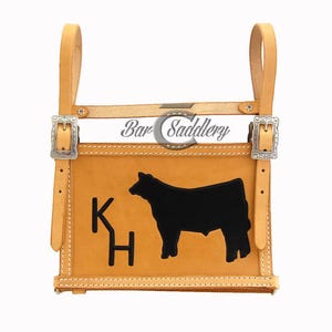 Può includere: Una borsa da sella in cuoio color cuoio con due cinghie verticali con fibbie argentate, cuciture bianche e una sagoma nera di una mucca accanto alle lettere KH. Il testo Bar Saddlery è stampato in nero sopra lo scomparto principale.