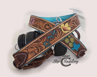 May include: A brown leather camera strap with turquoise accents and floral and cactus designs. The strap features a carved floral pattern, the letters "BS", and a Texas outline. The strap is paired with a black camera.