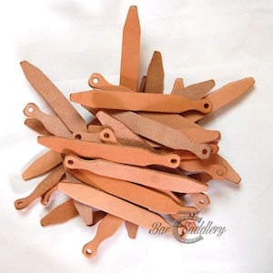 May include: A collection of light tan leather strips, each featuring a pointed end and a rounded end with a small circular hole. The leather pieces are stacked in a pile, and the text Bar Saddlery is visible in the bottom right corner.