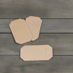 May include: Three light tan, rectangular leather pieces with clipped corners. Each piece features a smooth, flat surface and a uniform, natural beige color. The items are arranged with two overlapping at the top and one positioned horizontally below.