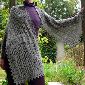 Puede incluir: Un chal de punto gris con un patrón de encaje. El chal está drapeado sobre los hombros de una persona y se lleva con una camisa morada.