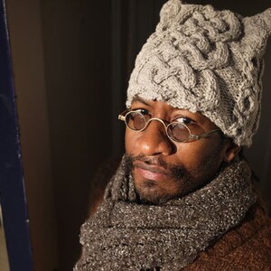 Puede incluir: Una persona con un gorro de punto gris con orejeras y gafas redondas. El gorro tiene un patrón trenzado y la persona lleva una chaqueta marrón.