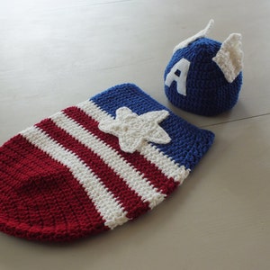 May include: A crocheted baby outfit in red, white, and blue, featuring a star and the letter 'A' on the hat. The outfit is designed to look like Captain America's costume.