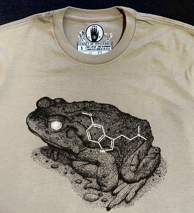 May include: A beige t-shirt with a black and white illustration of a toad with a chemical structure on its back. The shirt has a tag that says "Closet of Mysteries" and "S".