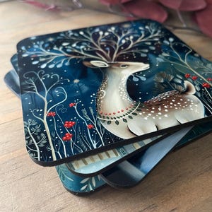 May include: A set of four coasters featuring a stylized illustration of a deer with antlers made of branches and leaves. The deer is standing in a forest setting with red berries and a dark blue background. The coasters are made of a durable material and are perfect for protecting your furniture from scratches and spills.