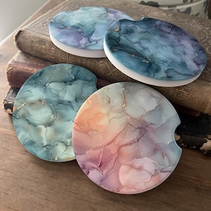May include: Four round coasters with abstract designs in shades of blue, green, pink, and purple with gold accents. The coasters are made of a white material and have a smooth surface.