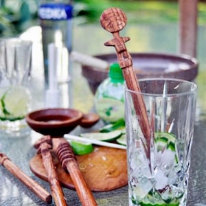 May include: A wooden cocktail muddler with a decorative top is in a glass with ice and lime slices. Other wooden muddlers and a wooden cutting board with lime slices are on the table. A bottle of vodka is in the background.