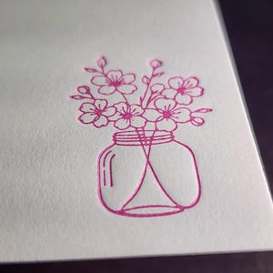 Puede incluir: Primer plano de una tarjeta blanca con un diseño floral rosa. El diseño presenta un tarro de albañil lleno de flores. Las flores están en plena floración y el tarro está detallado con líneas curvas.