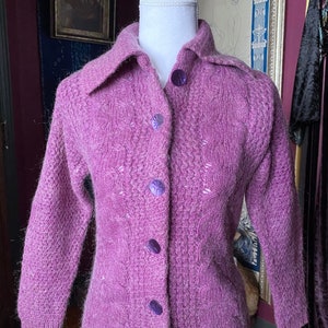 May include: A purple knitted cardigan with a cable knit pattern and a button-up front. The cardigan has a collar and long sleeves.