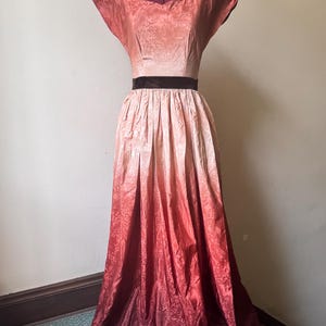 May include: A vintage floor-length dress in a gradient of colors from peach to deep red, displayed on a mannequin. The dress features a square neckline, short sleeves, and a dark brown velvet belt. The fabric has a subtle floral pattern.