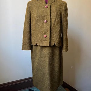 May include: A vintage olive green skirt suit. The jacket has a scalloped collar and three pink buttons. The matching skirt is knee-length. The suit is displayed on a dress form.
