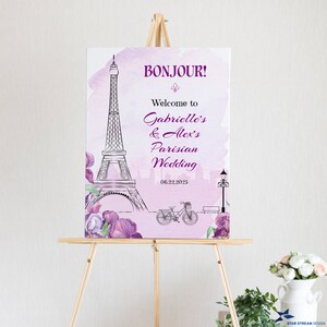 K&ouml;nnte beinhalten: Ein rosafarbenes und violettes Aquarell-Hochzeitschild mit einer Schwarzwei&szlig;illustration des Eiffelturms, eines Fahrrads und einer Stra&szlig;enlaterne. Der Text auf dem Schild lautet "Bonjour! Willkommen zur Pariser Hochzeit von Gabrielle und Alex 06.22.2025".