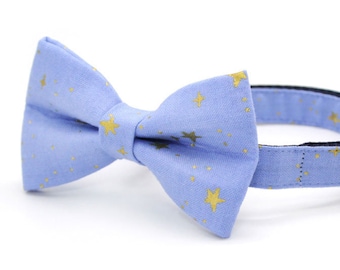 Puede incluir: Un collar para gato azul claro con un patrón de estrellas doradas y una pajarita a juego. El collar tiene una campanilla.