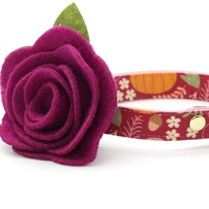 May include: A burgundy cat collar with a fall-themed pattern of pumpkins, acorns, and flowers. It features a large, deep pink felt rose with a green leaf accent. A gold bell and a black plastic buckle complete the design.