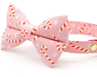 May include: Pink cat collar with a bow tie featuring a candy cane and snowflake pattern. The collar has a gold bell.