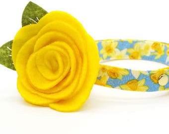 May include: A yellow felt flower with green leaves attached to a blue and yellow floral patterned cat collar with a bell.