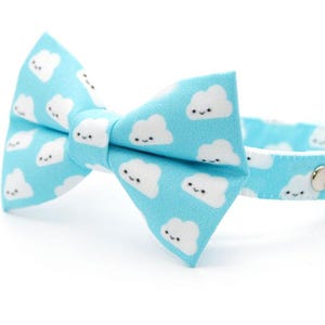 May include: A light blue cat collar with a matching bow tie. The collar and bow tie are patterned with white smiling cloud graphics. A small silver bell hangs from the collar.