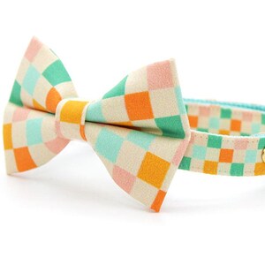 May include: A patterned cat collar and bow tie set. The bow tie and collar have a checkered design in orange, green, pink, and blue. A gold-coloured bell is attached to the collar. The collar is approximately 1.3 cm wide.