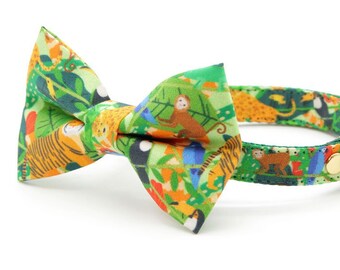 May include: A green cat collar with a bow tie featuring a jungle print with toucans, monkeys, tigers, and other animals. The collar has a gold bell.